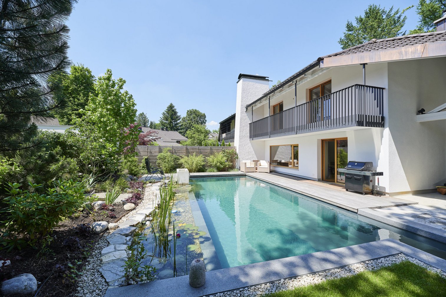 Modernes, zweistöckiges weißes Wohnhaus mit großem rechteckigem TeichMeister Swimmingpool im Garten, umgeben von Bäumen und Pflanzen, an einem sonnigen Tag.