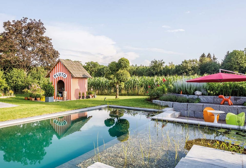 Garten mit Schwimmteich Ein TeichMeister Schwimmteich umgeben von grüner Wiese, Pflanzen und Bäumen, links ist ein Gartenhaus mit der Aufschrift „PLUTO“.