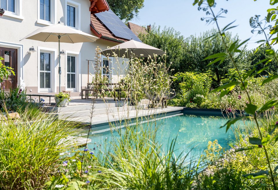 Naturgarten mit Pool neben einem hellen Wohnhaus mit Solaranlage; umgeben von wild-romantischer Bepflanzung, Sonnenschirmen und Sitzbereich auf der Terrasse.