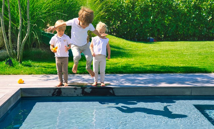 Drei kleine Kinder am Rand eines Gartenpools, eines springt lachend ins Wasser, während die anderen zusehen; im Hintergrund grüne Wiese und Hecke.