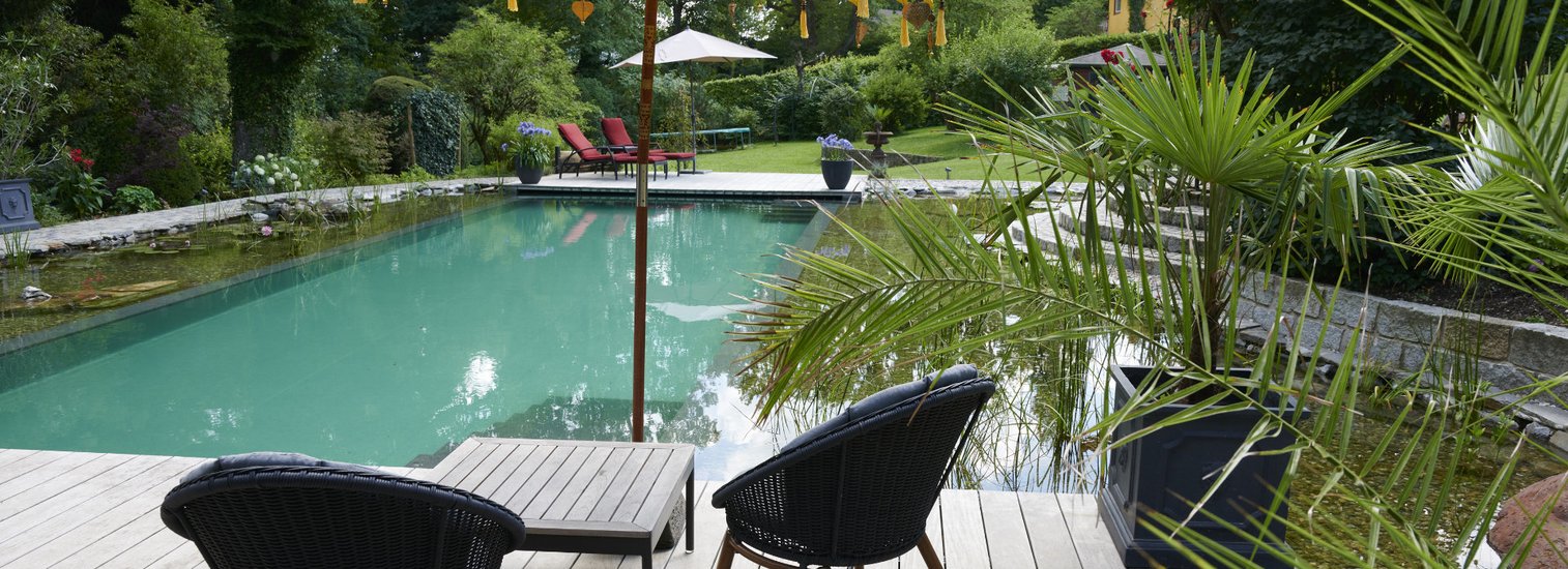 Ein ruhiger Garten mit einem TeichMeister Pool, zwei Stühlen und einem kleinen Tisch unter einem gelben Sonnenschirm auf einer Holzterrasse, umgeben von Bäumen und grünen Pflanzen.