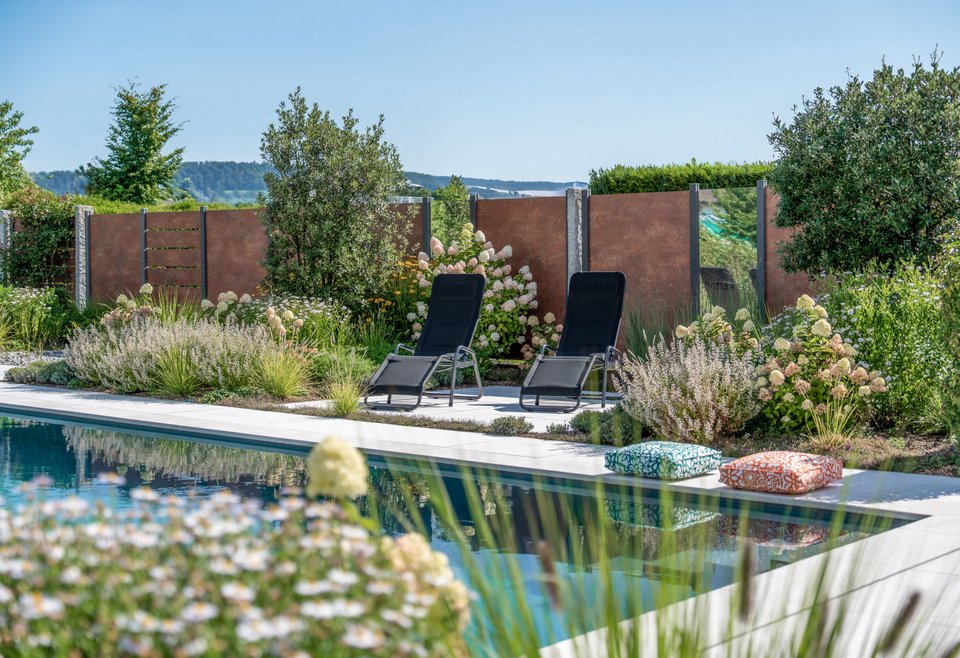 Naturpool Landschaft Moderner Garten mit rechteckigem Pool und zwei schwarzen Liegestühlen, umgeben von Blumen und Sträuchern, ruhige Atmosphäre an sonnigem Tag.