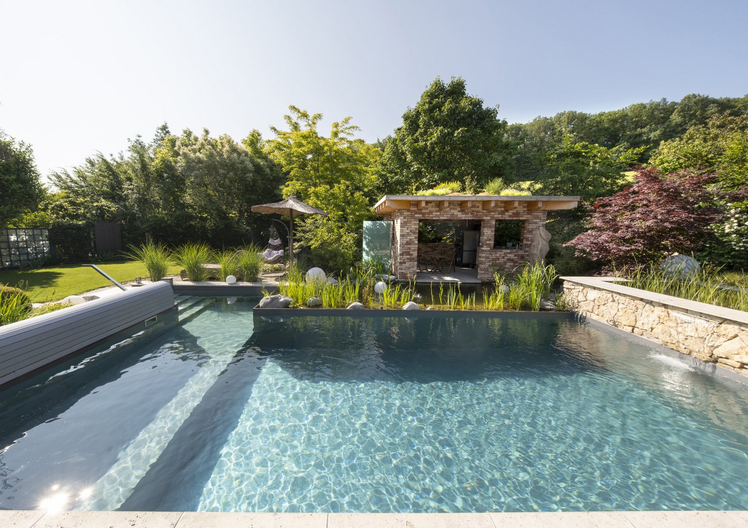 Ein klarer, moderner Swimmingpool in einem gepflegten Garten mit üppigen Pflanzen und Bäumen, daneben eine überdachte Lounge-Ecke aus Stein. Die Umgebung wirkt ruhig und einladend für Entspannung im Freien.