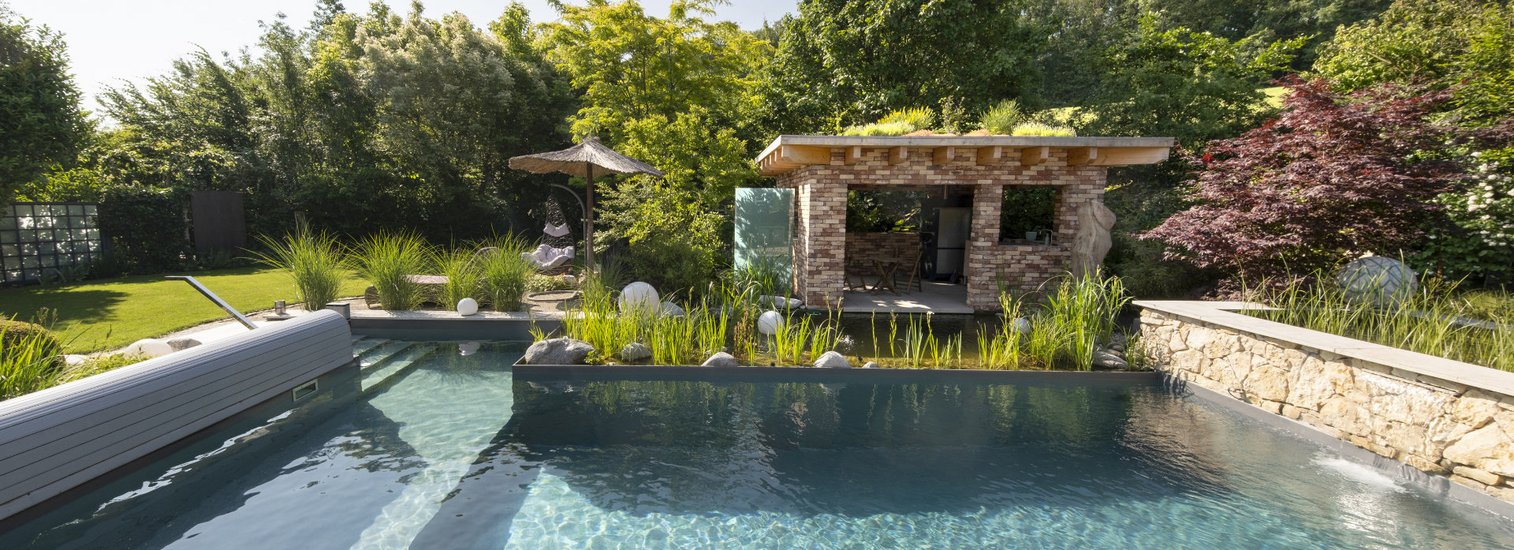 Ein klarer, moderner Swimmingpool in einem gepflegten Garten mit üppigen Pflanzen und Bäumen, daneben eine überdachte Lounge-Ecke aus Stein. Die Umgebung wirkt ruhig und einladend für Entspannung im Freien.
