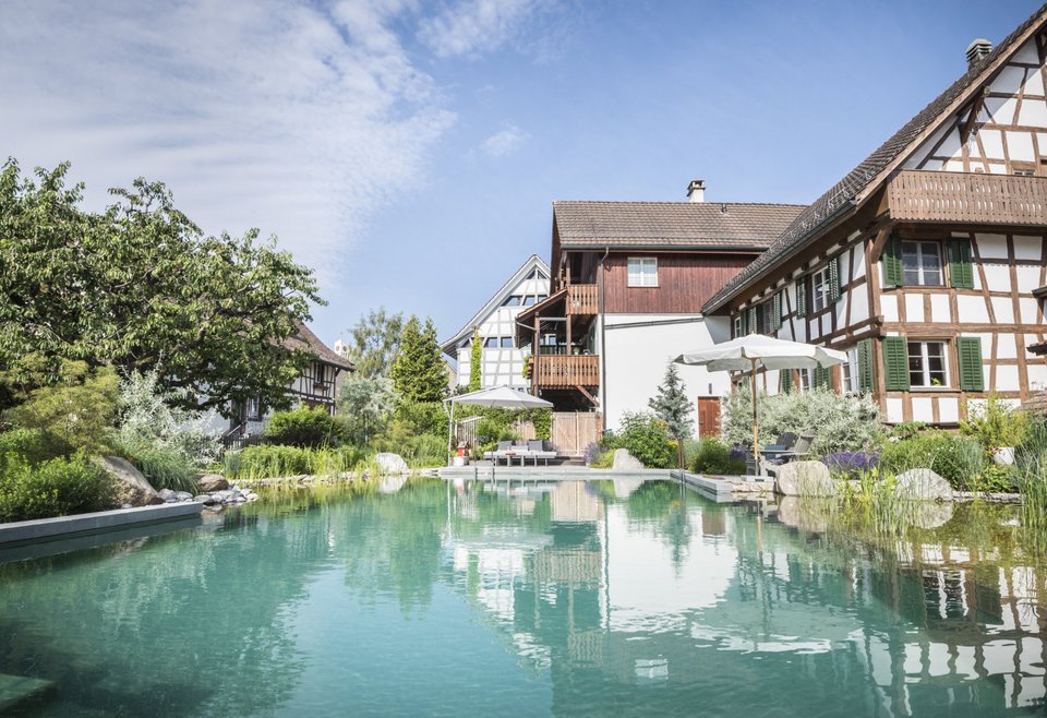 Naturpool Grimm Gärten Traditionelles Fachwerkhaus neben einem klaren Naturbadeteich mit Pflanzen und blauer Himmel.