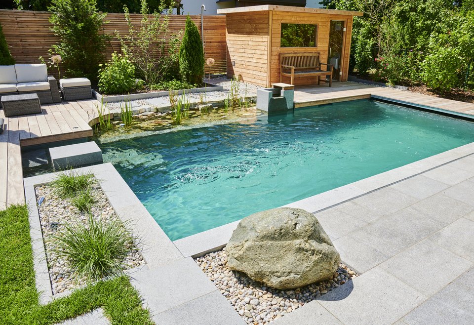 TeichMeister Schwimmteich mit klarem Wasser, umgeben von grauen Steinplatten, im Hintergrund eine kleine Holzsauna und Sitzgelegenheiten.