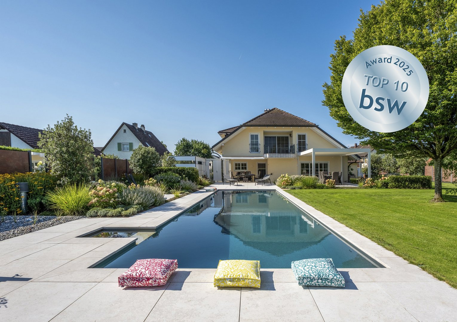 Pool mit Kissen Moderner Garten mit rechteckigem Swimmingpool vor einem Einfamilienhaus, umgeben von gepflegtem Rasen und Bäumen. Drei bunte Sitzkissen am Poolrand. Auszeichnung "Award 2025 TOP 10 bsw" als Symbol für Qualität und Design.