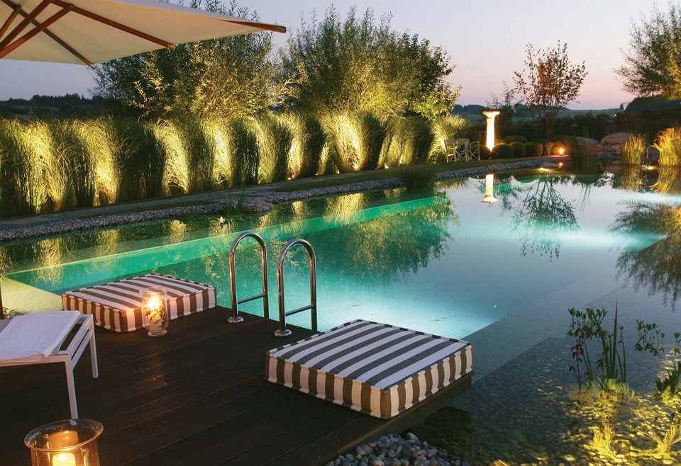 Schwimmteich mit Beleuchtung. Ein beleuchteter TeichMeister Schwimmteich bei Dämmerung, umgeben von Pflanzen und Bäumen, mit Liegen und Holzdeck.