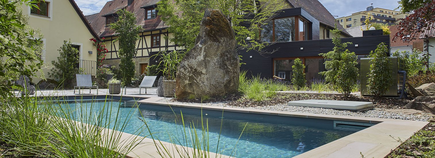 TeichMeister Naturpool in einem Garten mit Bepflanzung und großem Felsen in der Mitte, umgeben von traditionellen Fachwerkhäusern unter blauem Himmel.