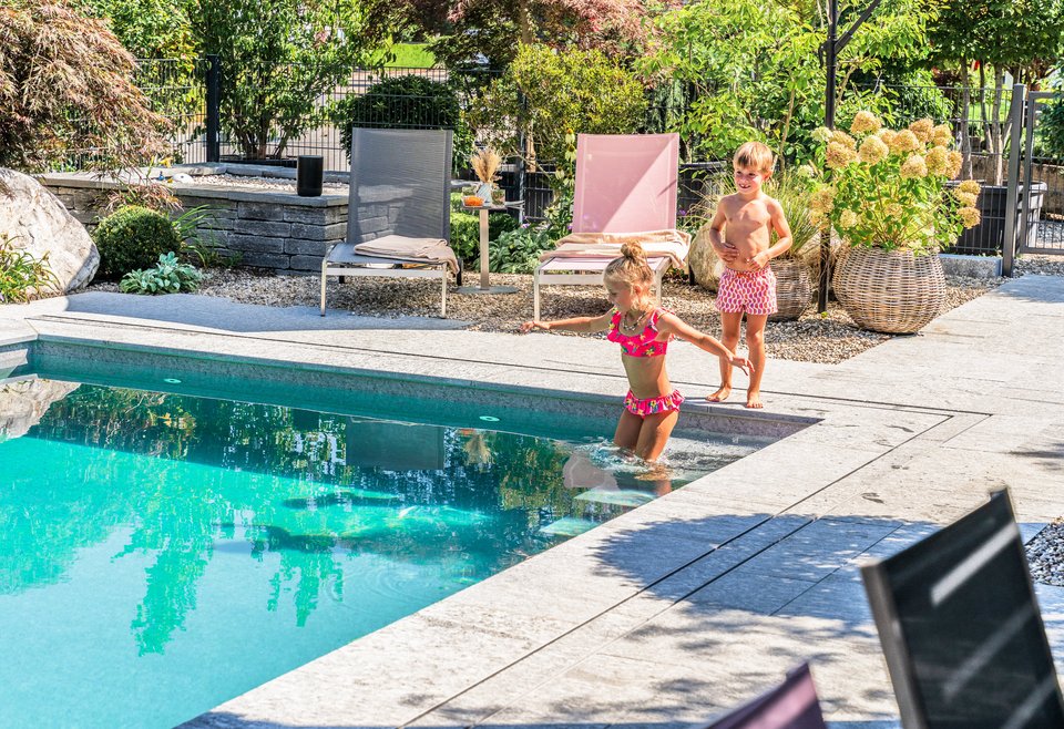 Kinder spielen im Naturpool Zwei kleine Kinder in Badesachen stehen an einem Poolrand in einem sonnigen Garten. Ein Mädchen steigt ins Wasser, ein Junge beobachtet sie. Im Hintergrund ruhige Gartenmöbel und grüne Pflanzen.