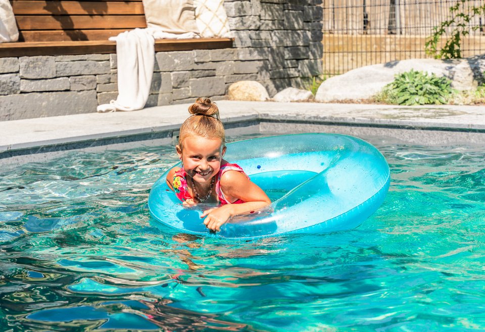 Kind im Schwimmring Ein junges Mädchen mit einem roten Schwimmring plantscht fröhlich in einem klaren Swimmingpool an einem sonnigen Tag.