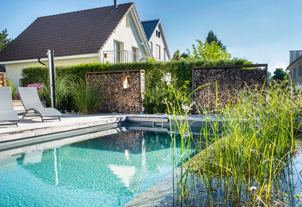 Schwimmteich mit Bepflanzung TeichMeister Schwimmteich mit klarem Wasser, eingebettet in einen gepflegten Garten mit hohen Gräsern und zwei Liegestühlen.