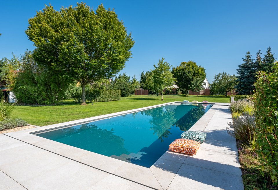 Swimmingpool rechteckig Moderner rechteckiger Swimmingpool in einem Garten mit grünem Rasen und großen Bäumen, bei klarem blauem Himmel. Am Poolrand liegen bunte Kissen zur Entspannung.