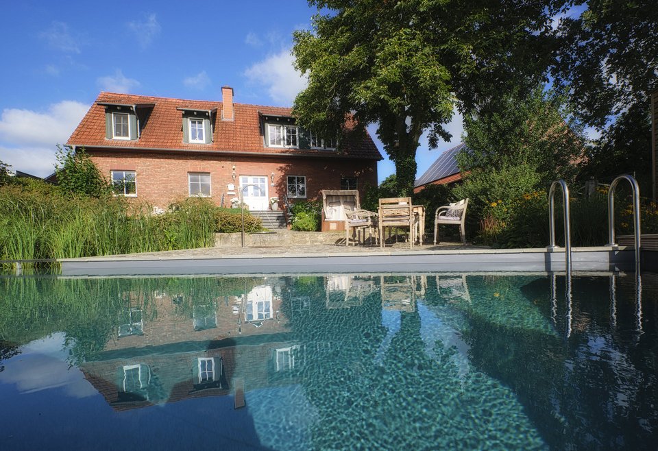 Ein rotes Backsteinhaus mit Dachfenstern und Terrasse, davor ein TeichMeister Swimmingpool, umgeben von Garten mit Bäumen und Pflanzen.