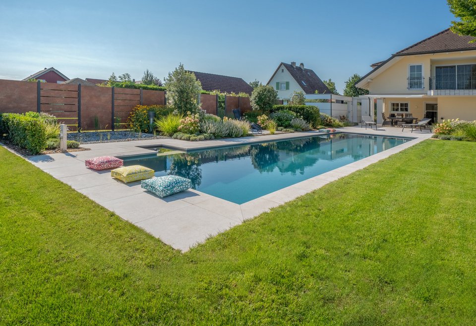 Offener Pool mit gepflegtem Garten Moderner rechteckiger Swimmingpool in einem gepflegten Garten mit Rasen und bunten Bodenkissen am Rand, angrenzend an ein großes, helles Wohnhaus.