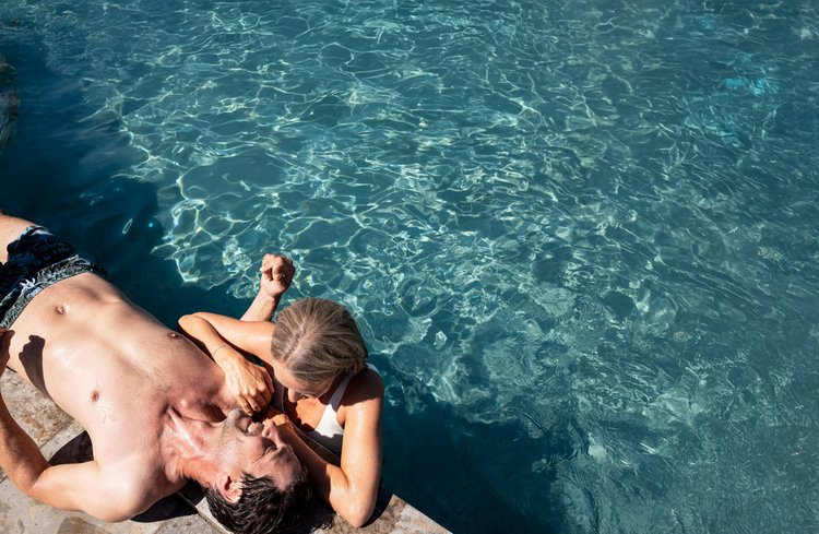 Ein Mann liegt am Beckenrand eines Pools, während eine Frau sich an ihn lehnt.