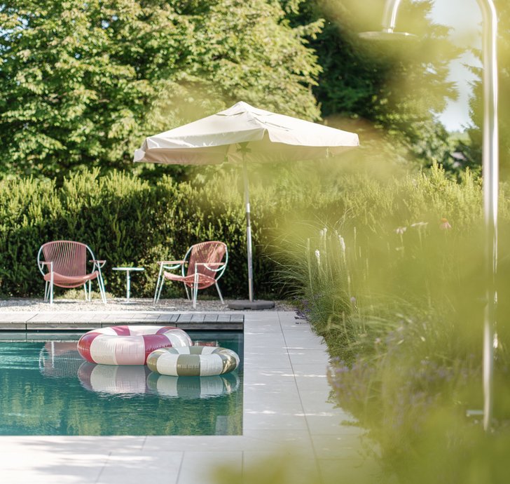 Moderner Gartenpool mit zwei bunten Schwimmringen, daneben zwei rosa Gartenstühle mit Beistelltisch und Sonnenschirm vor dichter Hecke.