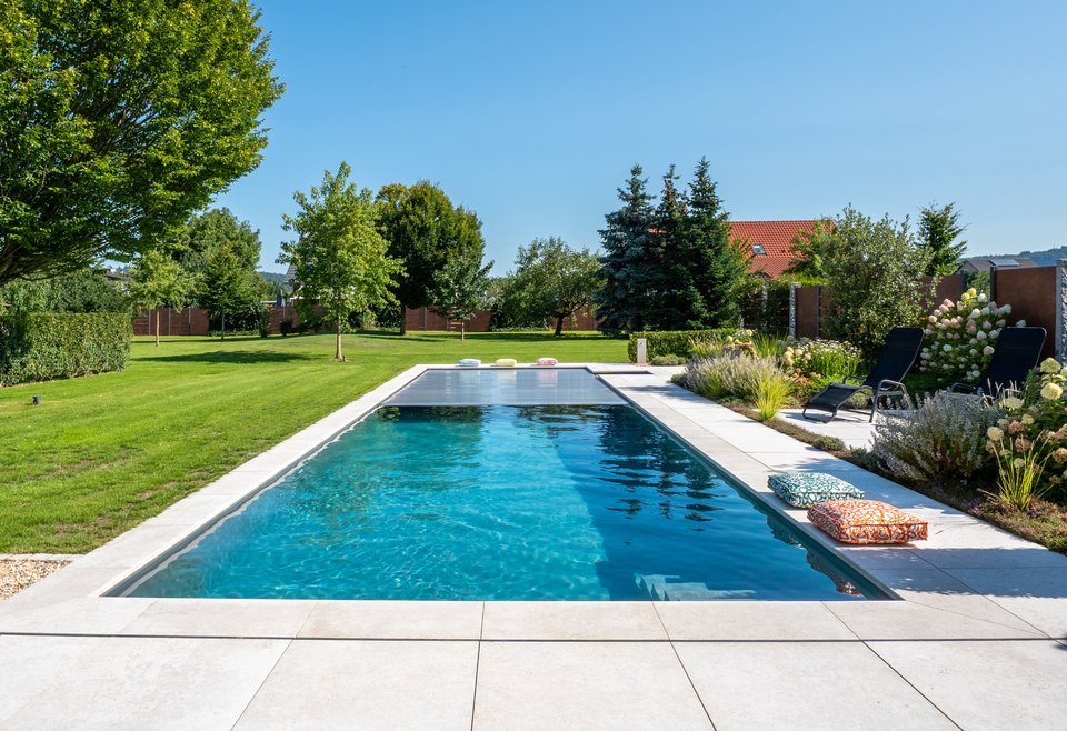 Pool Outdoor Moderner, rechteckiger Outdoor-Pool in einem gepflegten Garten mit grünem Rasen, Bäumen und Pflanzen. Am Beckenrand liegen bunte Kissen, daneben stehen zwei schwarze Liegestühle. Der Himmel ist klar und sonnig.