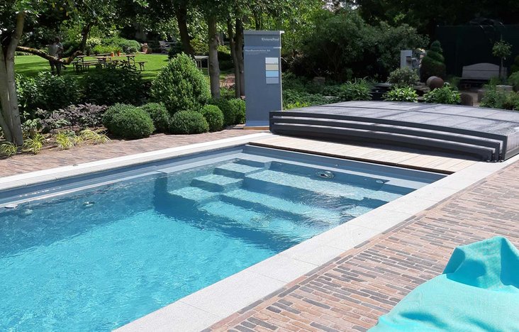 Ein moderner Pool in der TeichMeister Poolausstellung mit klar blauem Wasser und breiten Einstiegstreppen, umgeben von einer gepflasterten Liegefläche und Garten.