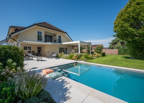 Moderner Hausgarten mit großem rechteckigem Swimmingpool, Sonnenterrasse mit Liegestühlen und Sitzbereich, gepflegtem Rasen und üppiger Bepflanzung, unter klarem blauem Himmel.