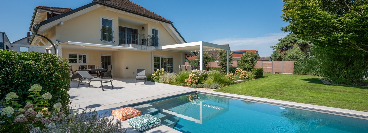 Moderner Hausgarten mit großem rechteckigem Swimmingpool, Sonnenterrasse mit Liegestühlen und Sitzbereich, gepflegtem Rasen und üppiger Bepflanzung, unter klarem blauem Himmel.
