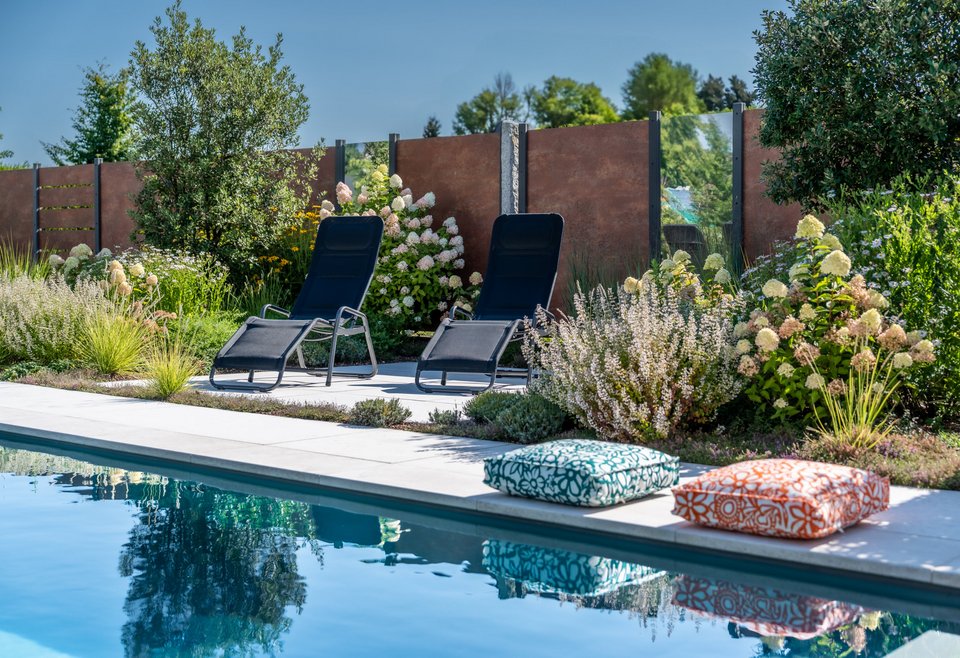 Poollandschaft Moderner Außenpool mit zwei schwarzen Liegestühlen und bunten Sitzkissen, umgeben von üppiger, blühender Bepflanzung und grünem Garten, unter klarem blauem Himmel.